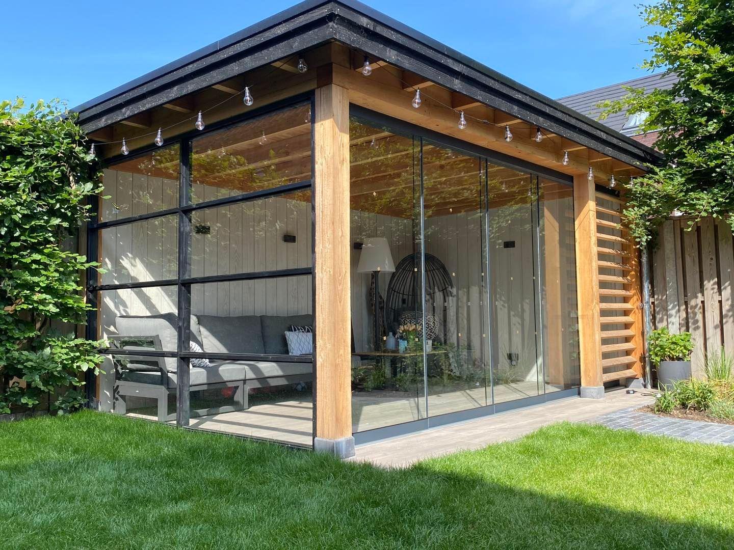 Modern glass enclosed outdoor seating area with wooden beams, string lights, and surrounded by greenery.