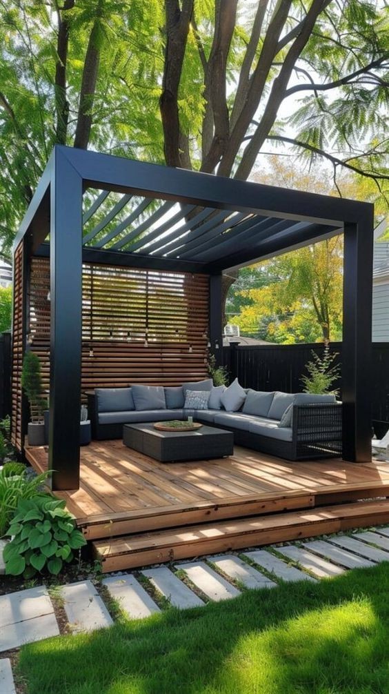 Modern outdoor pergola with a wooden deck, cushioned seating, and lush green surroundings.